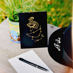 A matte black card with a zodiac illustration constellation design of Capricorn (stenbocken in Swedish), styled on a rustic wooden table with a white card backside, a black pen, and a plant in a blurred outdoor setting.